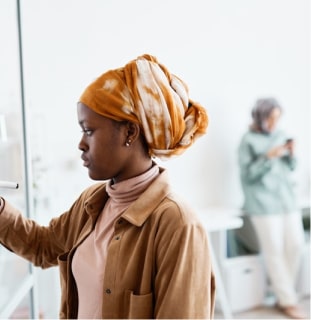 Person writing on whiteboard in office, another person on phone in background.