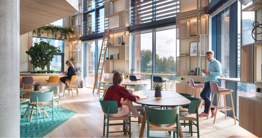 Modern coworking space with people, natural light, greenery, and stylish furniture.